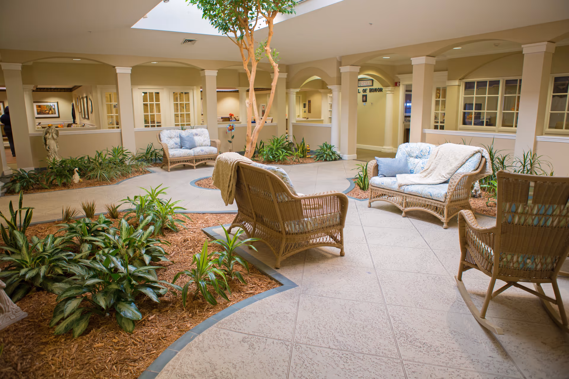 Indoor courtyard-style seating area with wicker chairs, sofas, plants, and a skylight in a senior living facility.