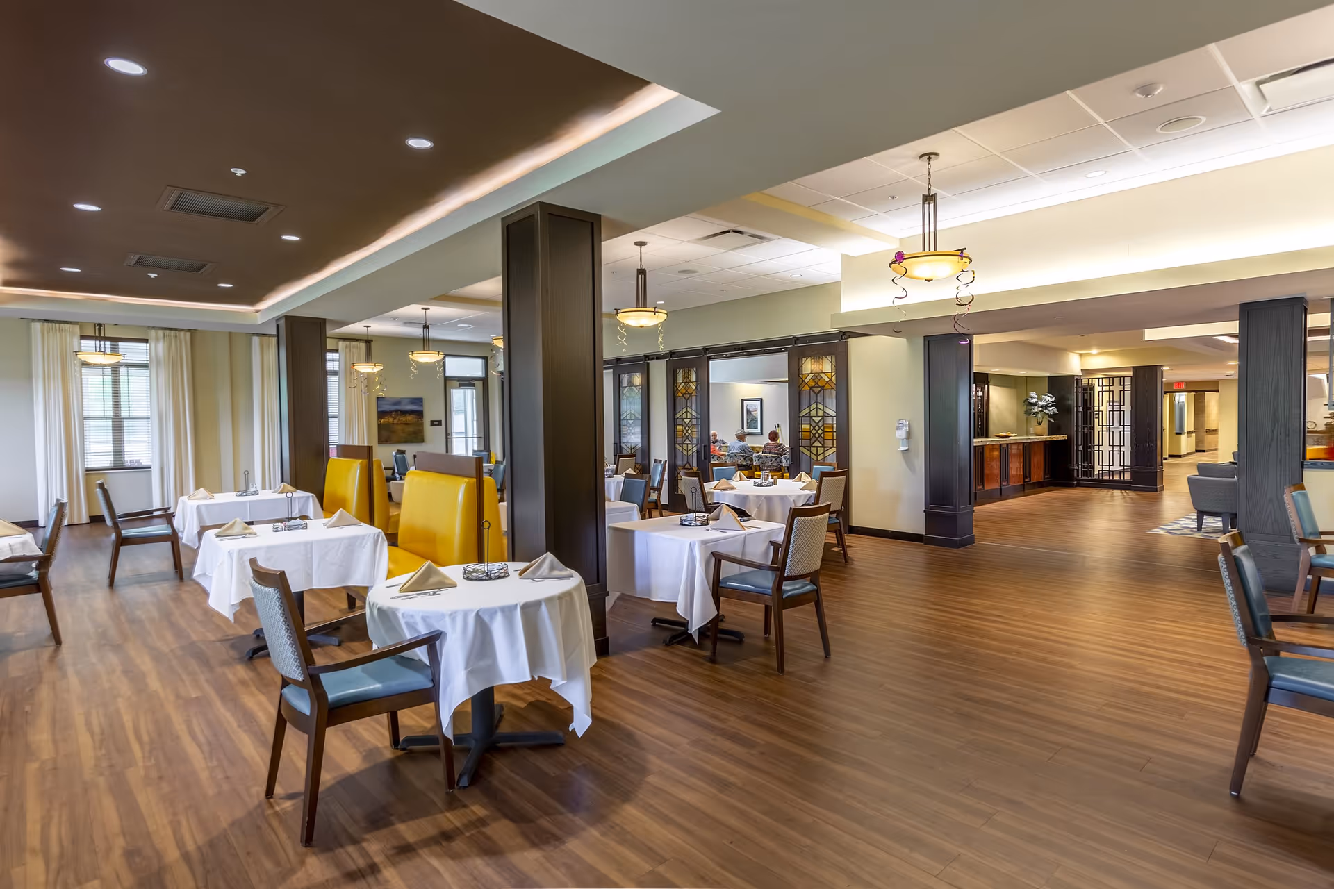 A spacious dining room in a senior living facility with several round tables covered with white tablecloths, each set with napkins and chairs. The room features wooden flooring, yellow cushioned booth seating, large windows with curtains, and warm overhead lighting fixtures. In the background, a few people are seated at a table in a separate dining area.