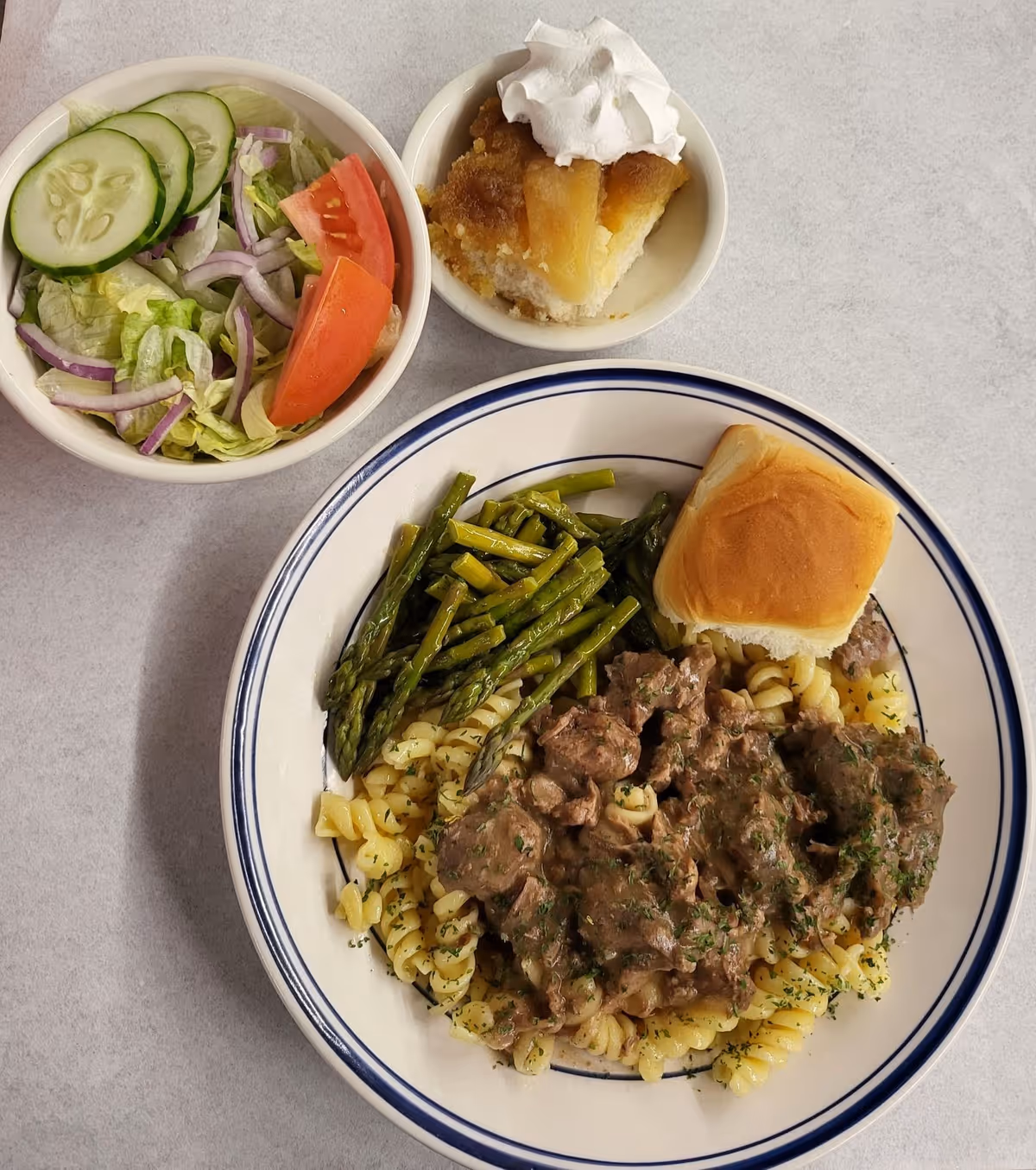 Top-down view of a plated meal featuring rotini pasta with beef, asparagus and a dinner roll, accompanied by a side salad and a dessert cup with whipped cream on a gray table.