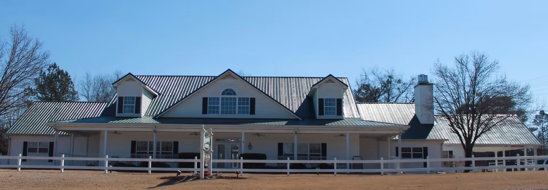 Wide exterior view of a single-story white building with a green metal roof, featuring dormer windows and a chimney. The building is surrounded by a white fence and leafless trees under a clear blue sky.