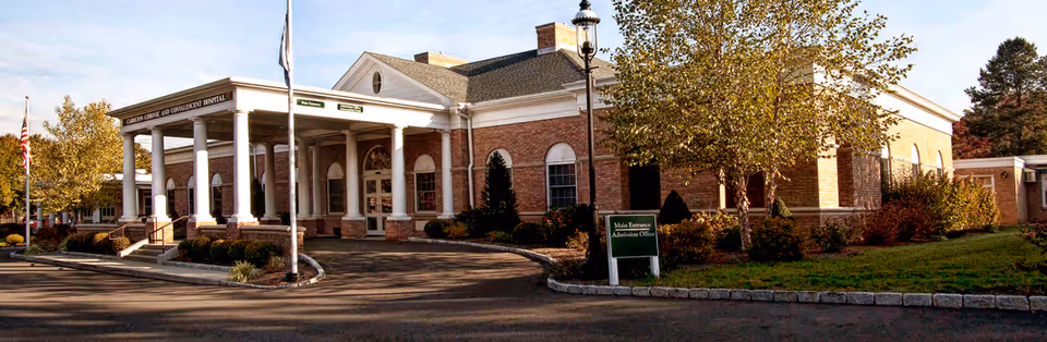 Exterior view of Carolton Chronic Convalescent Hospital building with brick walls, white columns at the entrance, a flagpole with an American flag, trees with autumn foliage, and a sign indicating the main entrance and admission office.