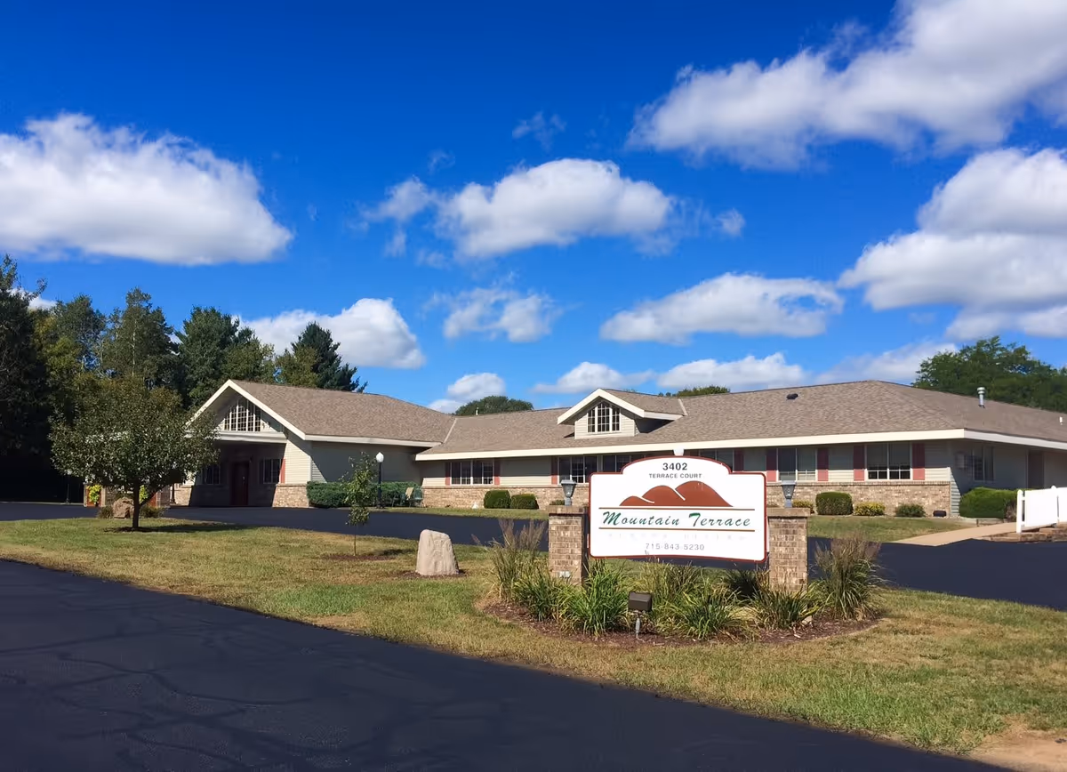 Exterior view of Mountain Terrace Senior Living building under a blue sky with scattered clouds. The building has a beige and brick facade with a brown roof. In front, there is a sign displaying the facility name and contact number, surrounded by grass, plants, and a paved driveway.
