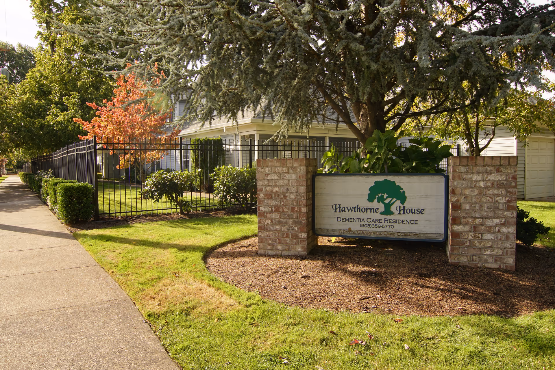Brick entrance sign for Hawthorne House Dementia Care Residence in front of a fenced yard and house.