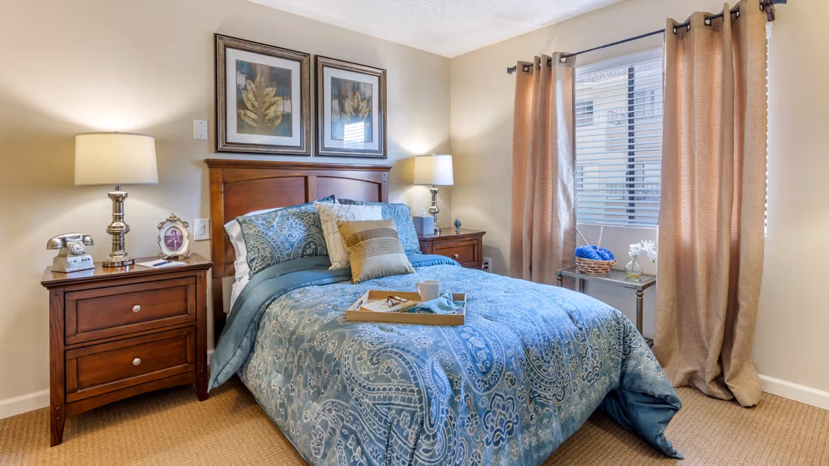 A neatly decorated bedroom with a made bed featuring blue patterned bedding, wooden nightstands and lamps, framed art above the headboard, and a window with beige curtains.