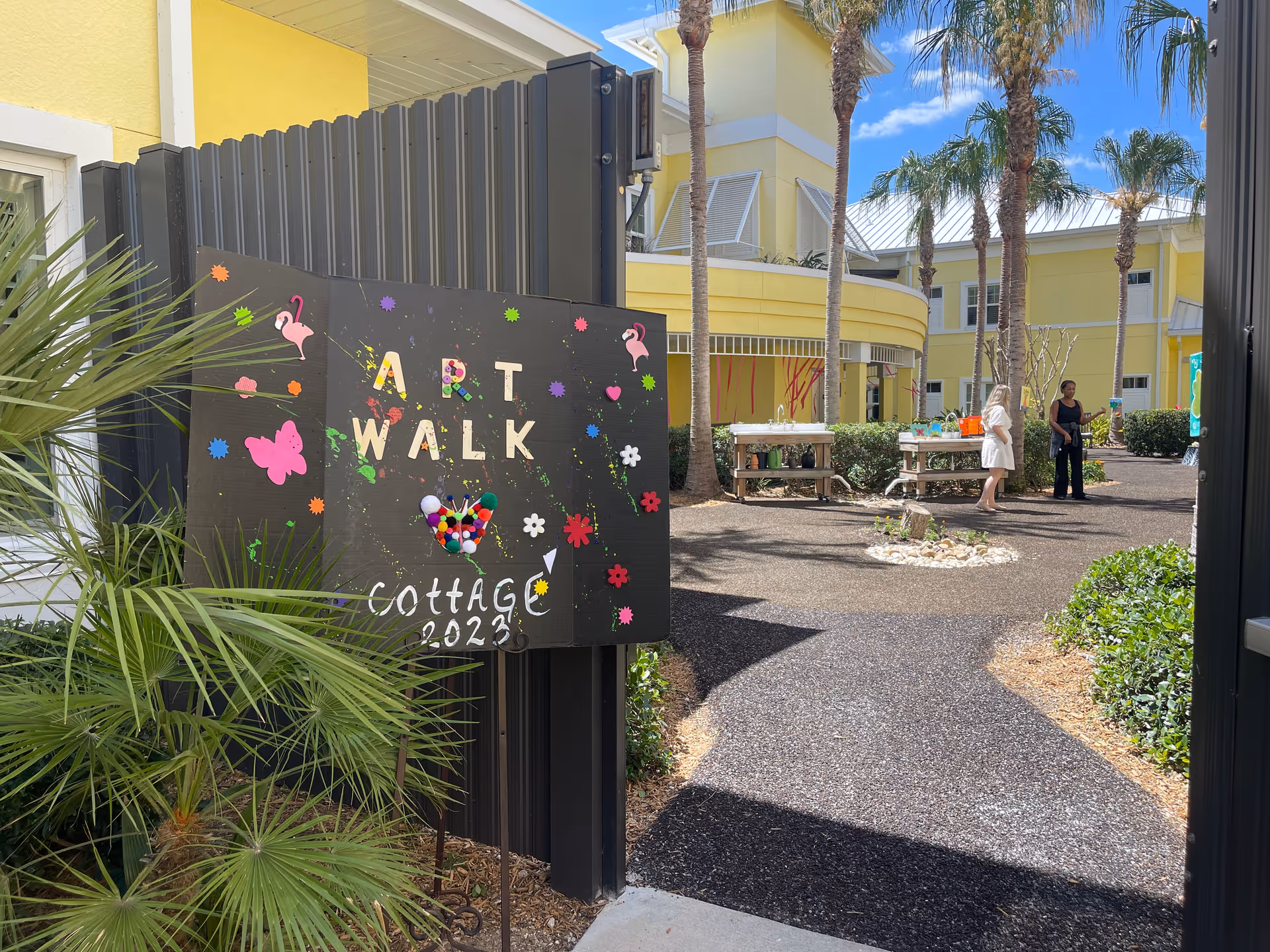 Outdoor courtyard area at The Cottage At Cypress Cove with a decorative sign reading 'ART WALK Cottage 2023' featuring colorful shapes and pom-poms. The courtyard has palm trees, bushes, and two women standing near tables with art supplies under a sunny blue sky.
