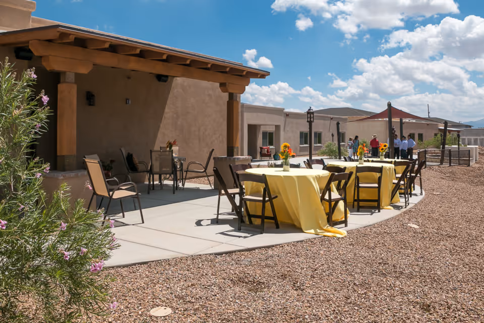 Outdoor patio area with tables covered in yellow tablecloths and decorated with vases of sunflowers. Several chairs are arranged around the tables. In the background, a group of people is standing near a building with a covered porch. The sky is partly cloudy.