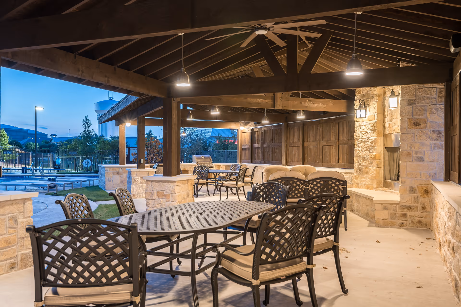 Covered outdoor patio area with metal tables and chairs, cushioned seating, stone pillars, and a stone fireplace. The patio is lit with hanging lights and overlooks a fenced pool area with trees and buildings in the background during dusk.