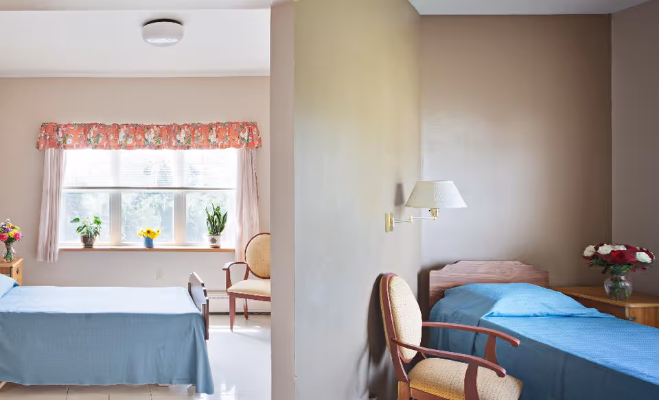 A bright room with two single beds separated by a partial wall. Each bed has a blue bedspread. There are chairs next to each bed, a window with floral curtains and potted plants on the windowsill, and a wall-mounted lamp above one bed. A vase with flowers is on a nightstand beside one bed.