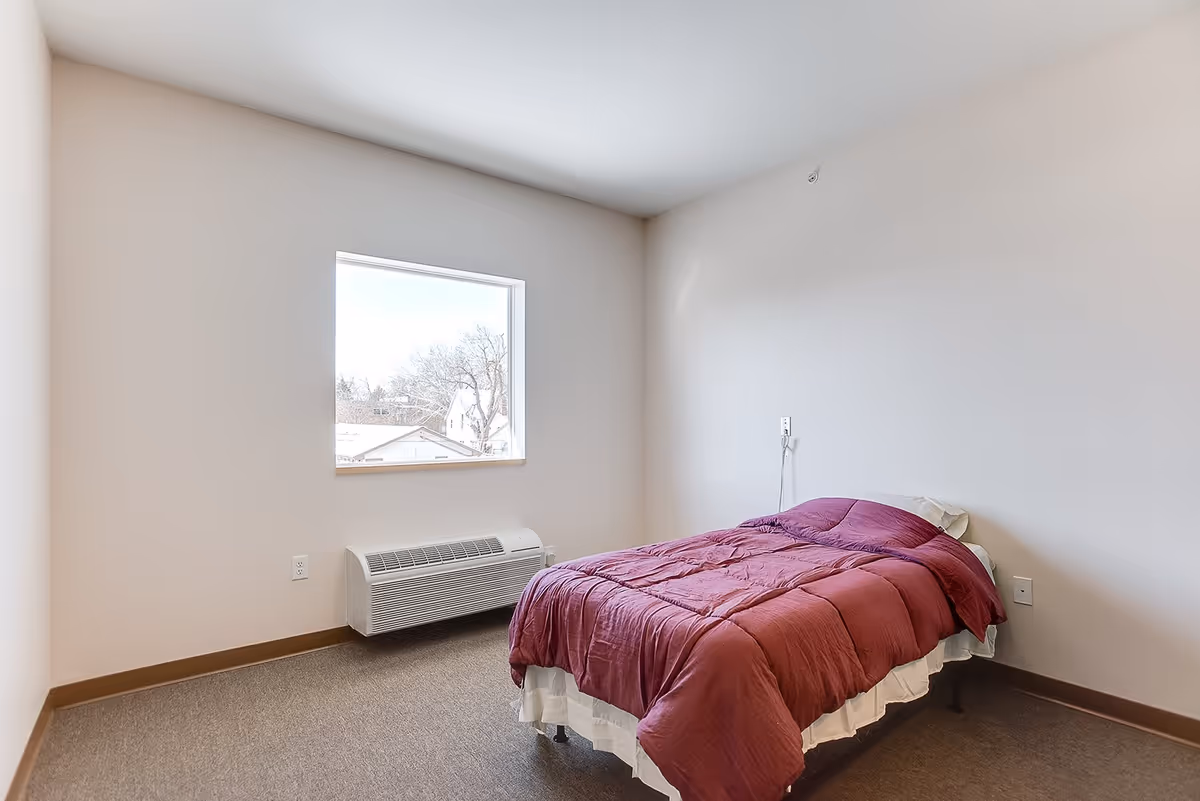 Spare bedroom with a single bed covered in a red comforter, a window, and a wall-mounted heater.