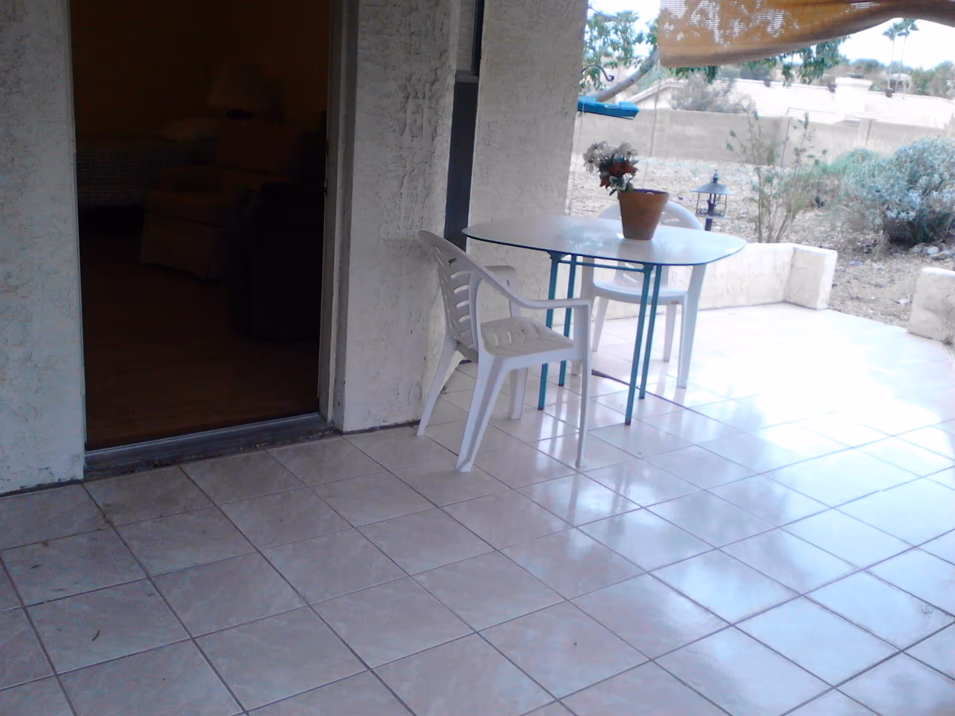 Outdoor tiled patio area with a small glass table and two white plastic chairs. A potted plant with flowers is on the table. The patio is adjacent to a building with a textured wall and an open door leading inside, where a lamp and furniture are partially visible. In the background, there is a garden area with bushes and a low wall.