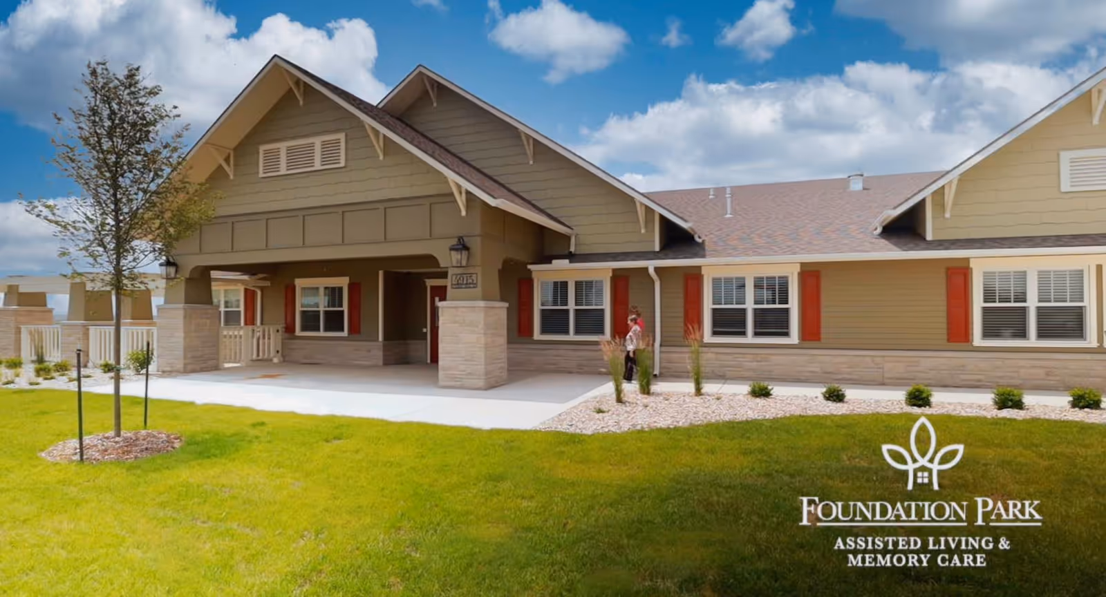 Exterior view of Foundation Park Assisted & Memory Care facility showing a single-story building with a covered entrance, beige siding, red window shutters, and a well-maintained lawn with a small tree and landscaping under a partly cloudy sky.