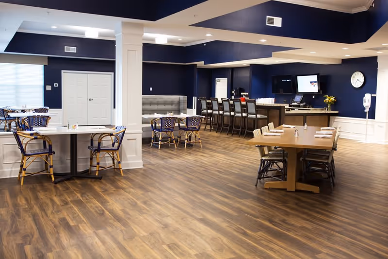 A spacious dining area with wooden flooring and navy blue walls. The room features multiple tables with chairs, including a long table set with napkins and salt and pepper shakers, a counter with bar stools, and a booth seating area. There are two wall-mounted screens and a clock on the wall, along with a hand sanitizer dispenser.