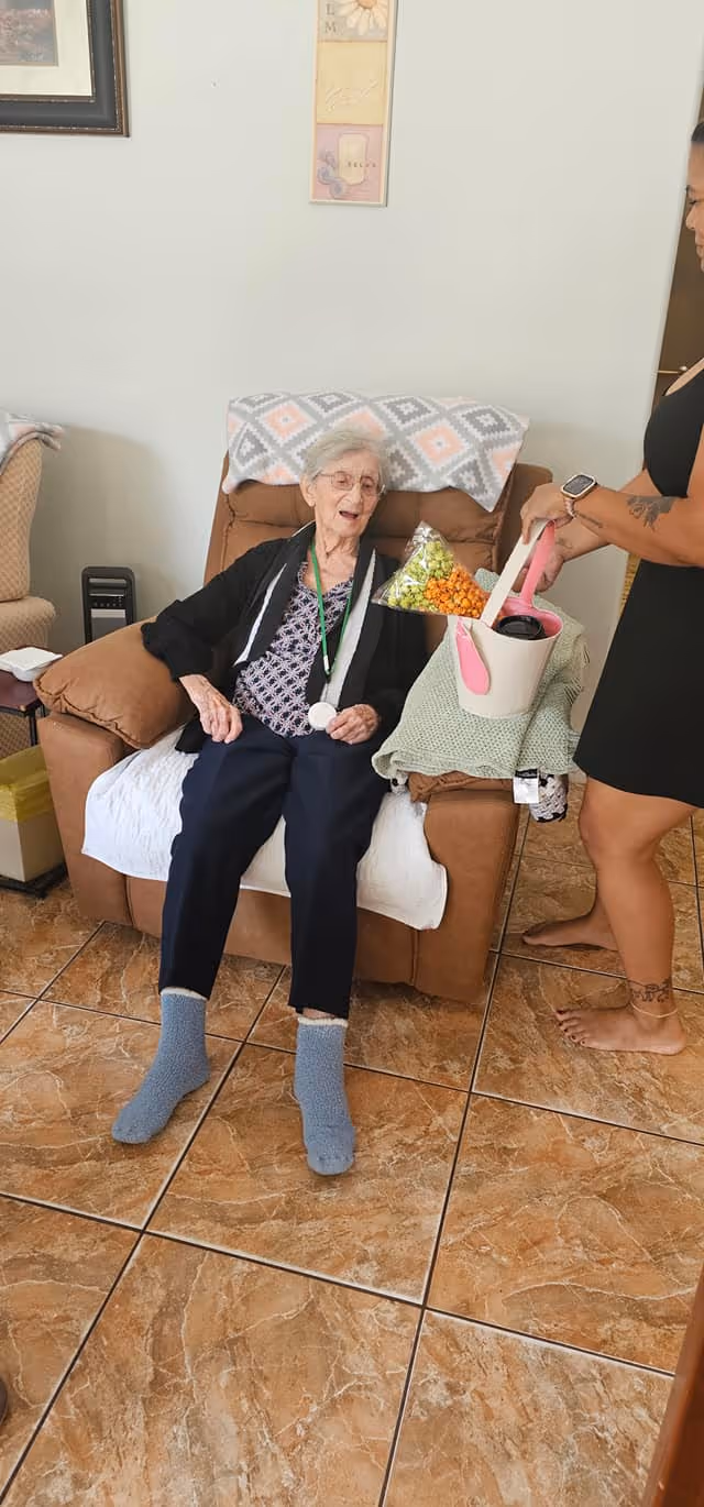 An elderly woman sitting comfortably in a brown recliner chair with a patterned blanket draped over the back. She is wearing glasses, a black cardigan, a patterned blouse, dark pants, and blue socks. A younger woman standing nearby is holding a white and pink basket with a handle, which contains a drink and some snacks. The room has tiled flooring and light-colored walls with framed artwork.