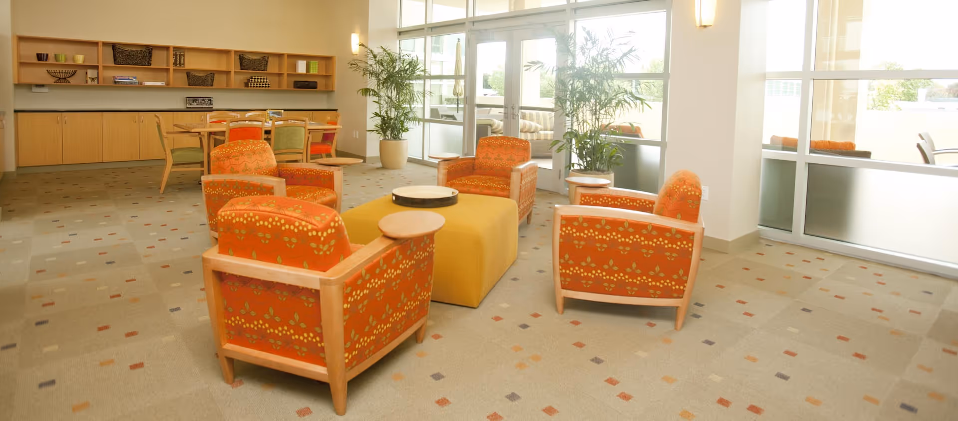 A bright and spacious common area with four orange patterned armchairs arranged around a yellow ottoman. There are large windows letting in natural light, potted plants near the windows, and a wooden cabinet with shelves and a dining table with chairs in the background.