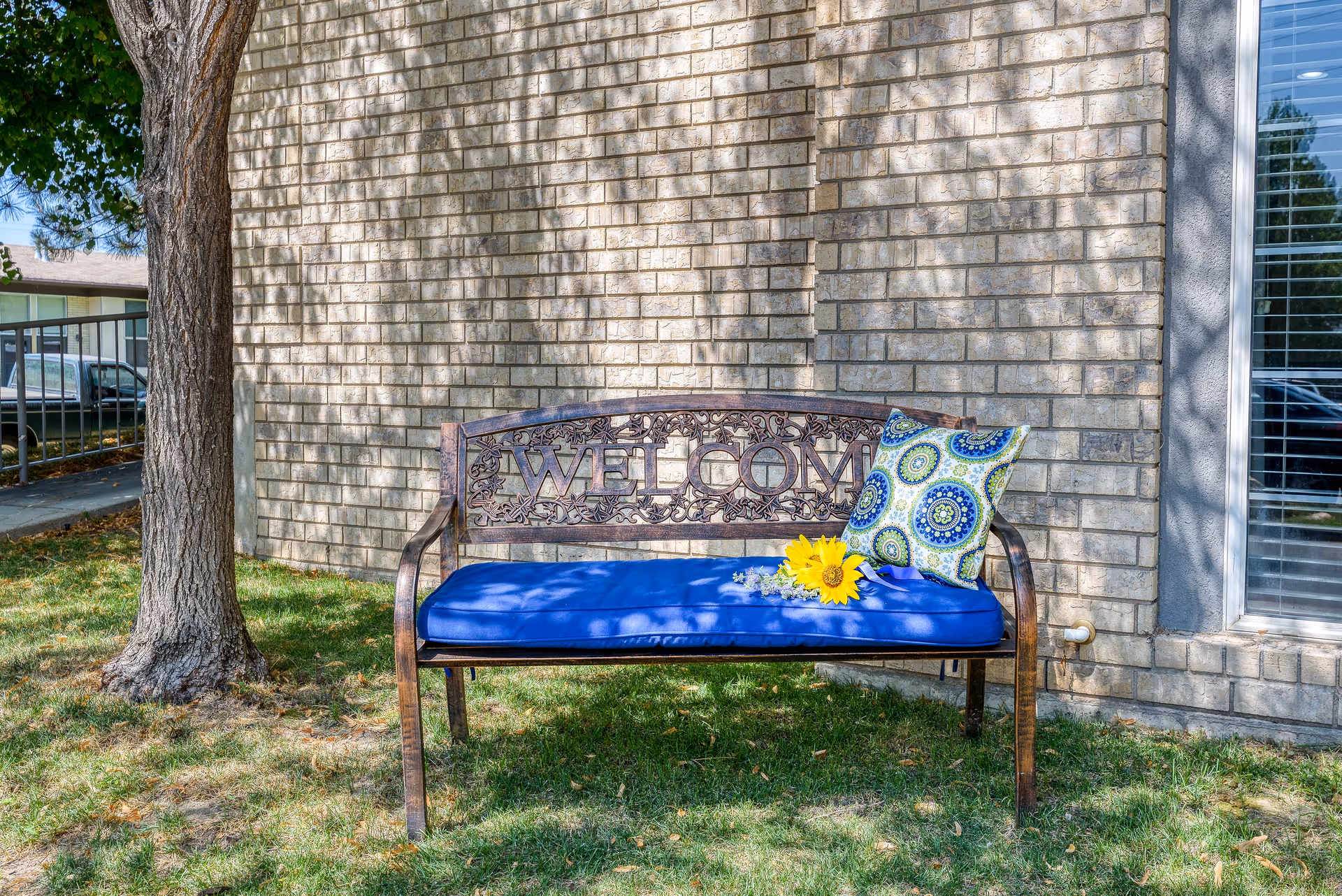 A decorative metal bench with the word 'WELCOME' on the backrest sits on grass next to a tree and a brick wall. The bench has a blue cushion and a patterned pillow with blue and green circular designs. A sunflower and small purple flowers rest on the cushion. A window with blinds is visible on the right side of the image.
