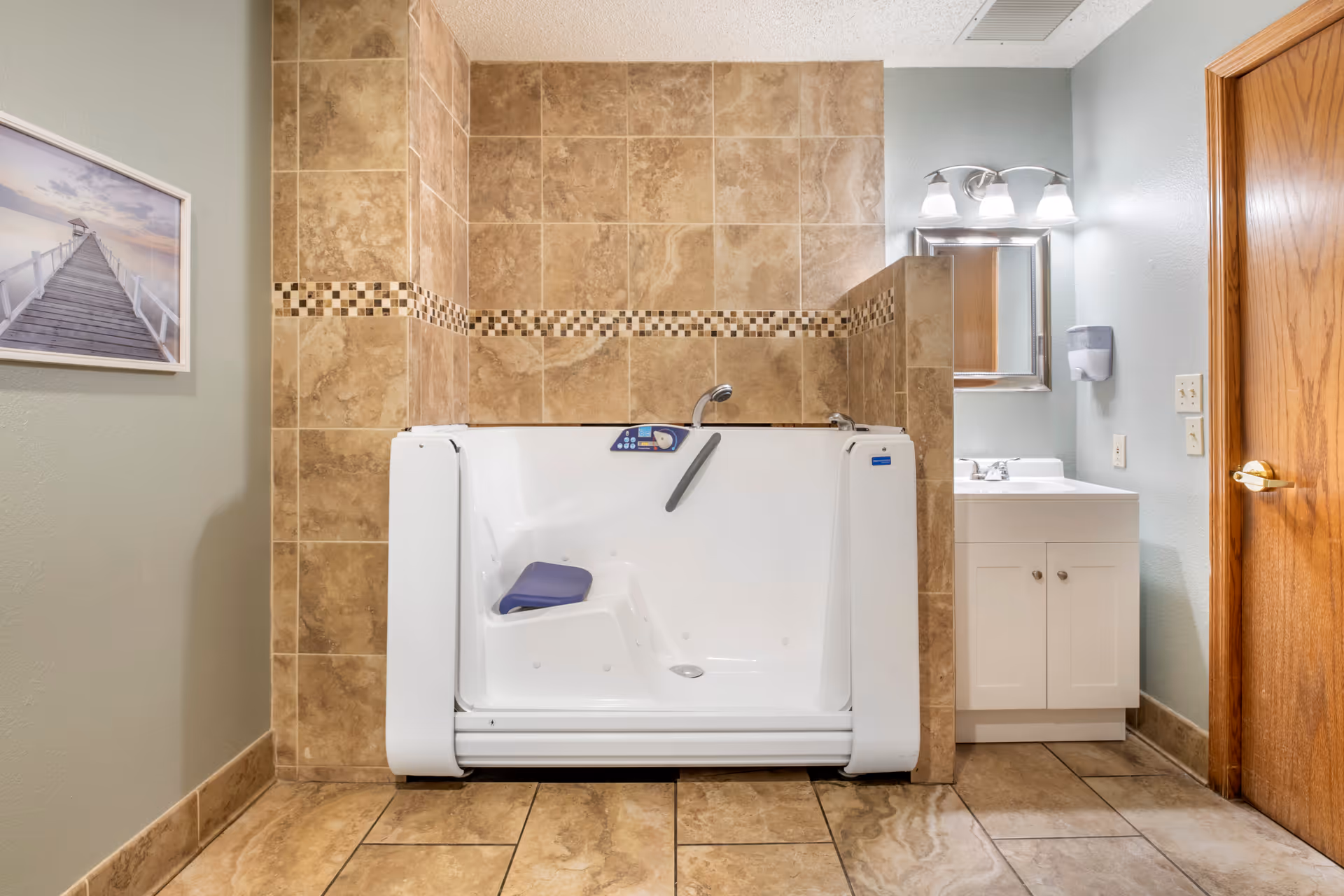 A bathroom with beige tiled walls and floor featuring a white walk-in bathtub with a blue seat and control panel. To the right is a white sink with a mirror and three light fixtures above it, and a wooden door. On the left wall, there is a framed picture of a pier extending into the water at sunset.