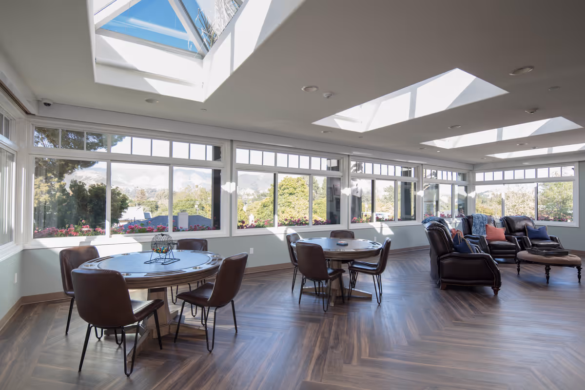 Bright sunlit common room with round game tables, leather chairs and sofas beneath skylights and large windows overlooking trees.