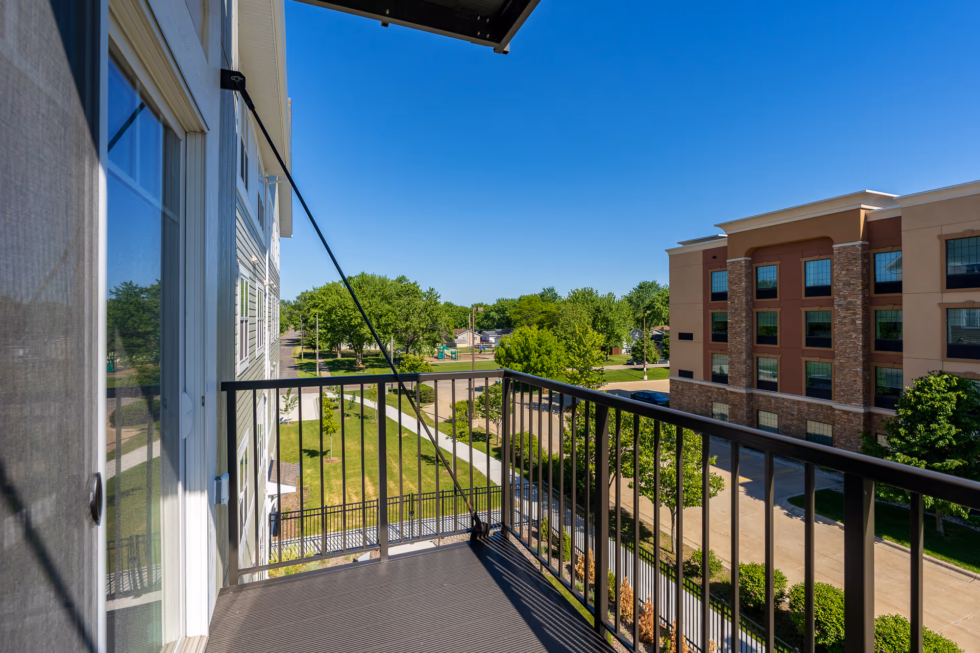 View from a balcony with a metal railing overlooking a landscaped courtyard, trees, and an adjacent multi-story building under a clear blue sky.