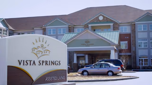 Exterior view of the Vista Springs assisted living facility building with a sign in the foreground displaying the facility name and logo. Several cars are parked in front of the entrance under a covered driveway.