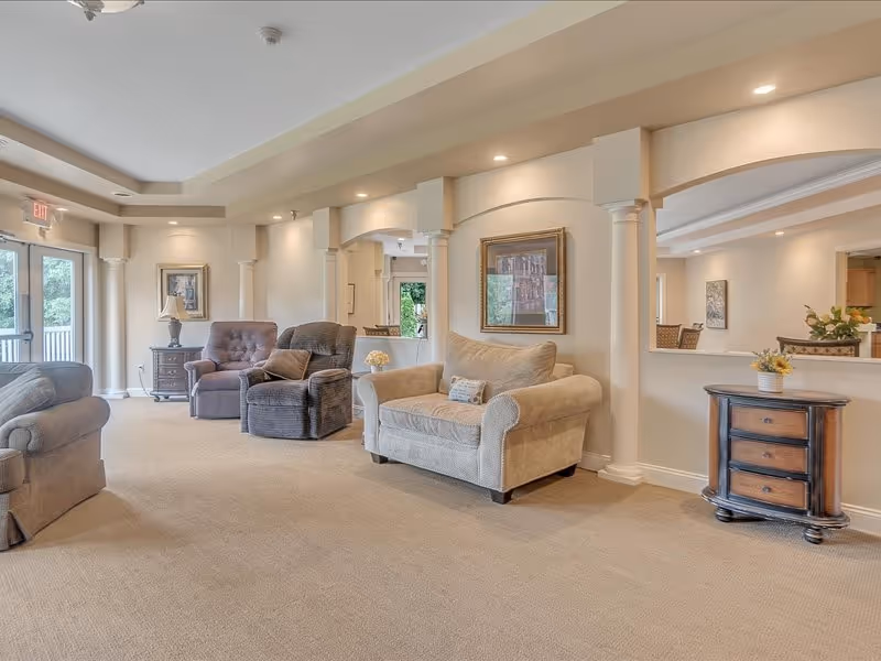 Sunlit senior living lounge with upholstered armchairs and a loveseat, decorative columns, side tables and framed artwork.