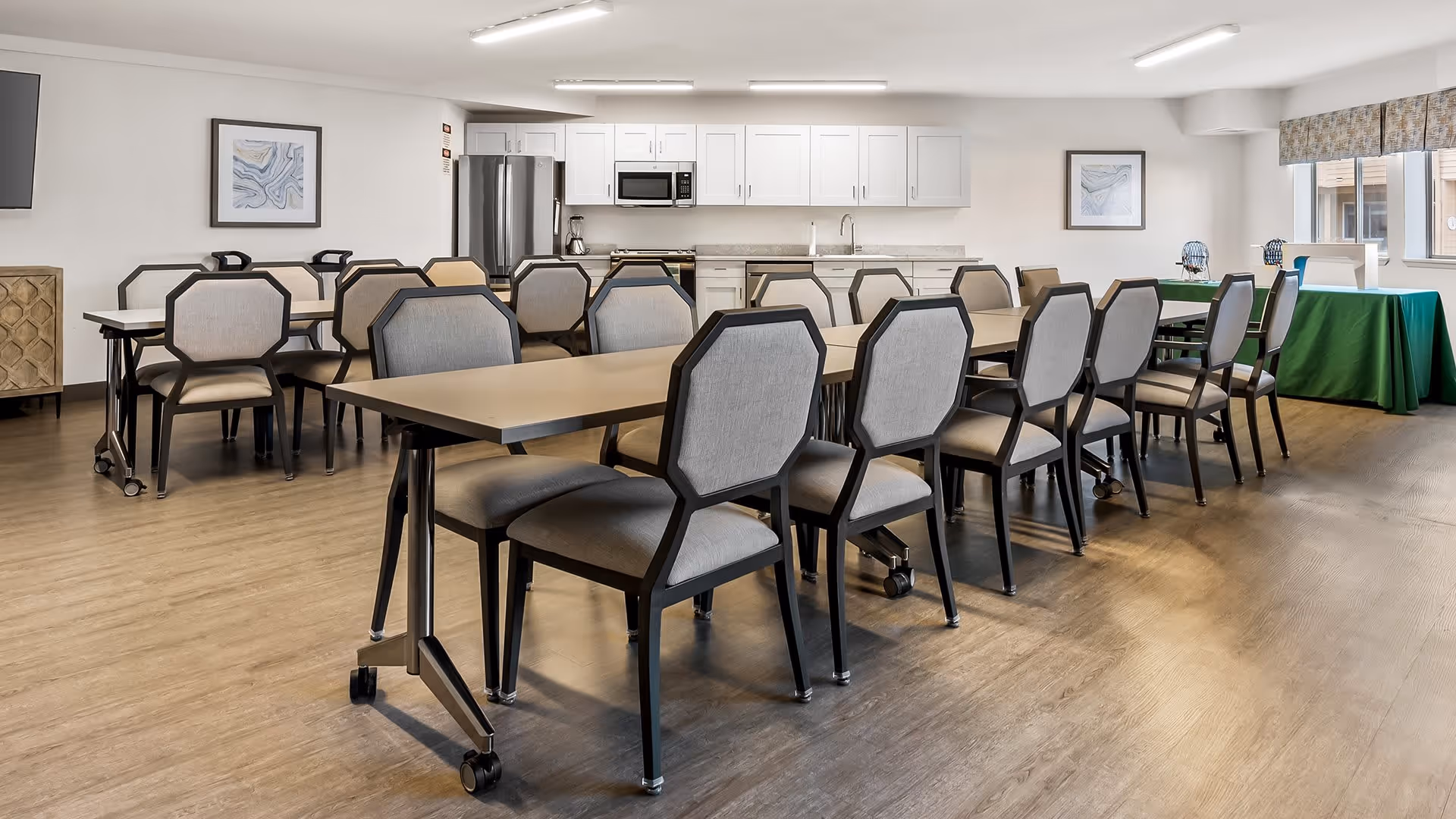 A spacious dining room with multiple tables and cushioned chairs arranged neatly. The room features a kitchenette area with white cabinets, a stainless steel refrigerator, microwave, and coffee maker. There are framed artworks on the walls and large windows with patterned valances letting in natural light. The floor is wood-style, and the overall ambiance is clean and welcoming.
