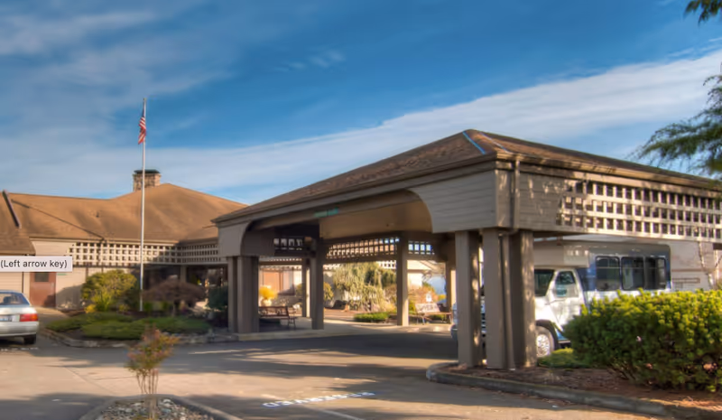 Exterior view of Channel Point Village facility entrance with a covered drop-off area, an American flag on a flagpole, a parked car, a white shuttle bus, and landscaped greenery under a blue sky with some clouds.