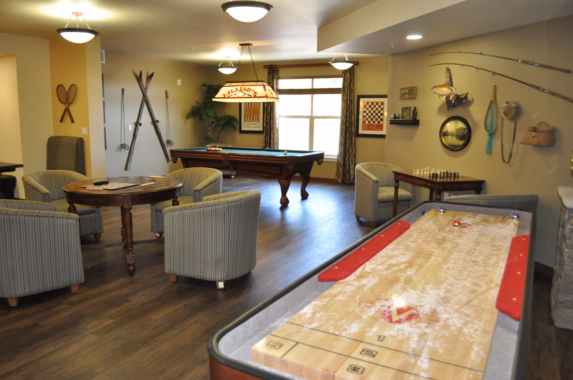 A senior living recreation room with a shuffleboard table in the foreground, a pool table and round card table surrounded by chairs, and fishing-themed wall decor.