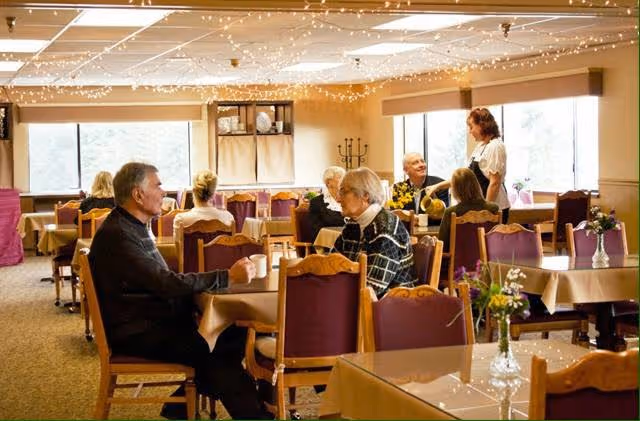 A dining room in a senior living facility with several elderly people sitting at tables covered with beige tablecloths. The room is warmly lit with string lights hanging from the ceiling. There are large windows letting in natural light, and a staff member is standing and talking to some of the residents. Small flower arrangements are placed on the tables.