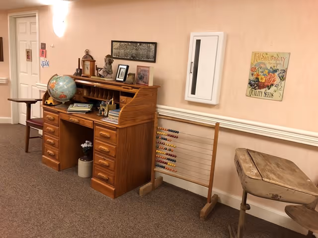 A wooden roll-top desk with a globe, books and decorative items against a pale wall in a hallway, with an abacus and an old wooden school desk beside it.