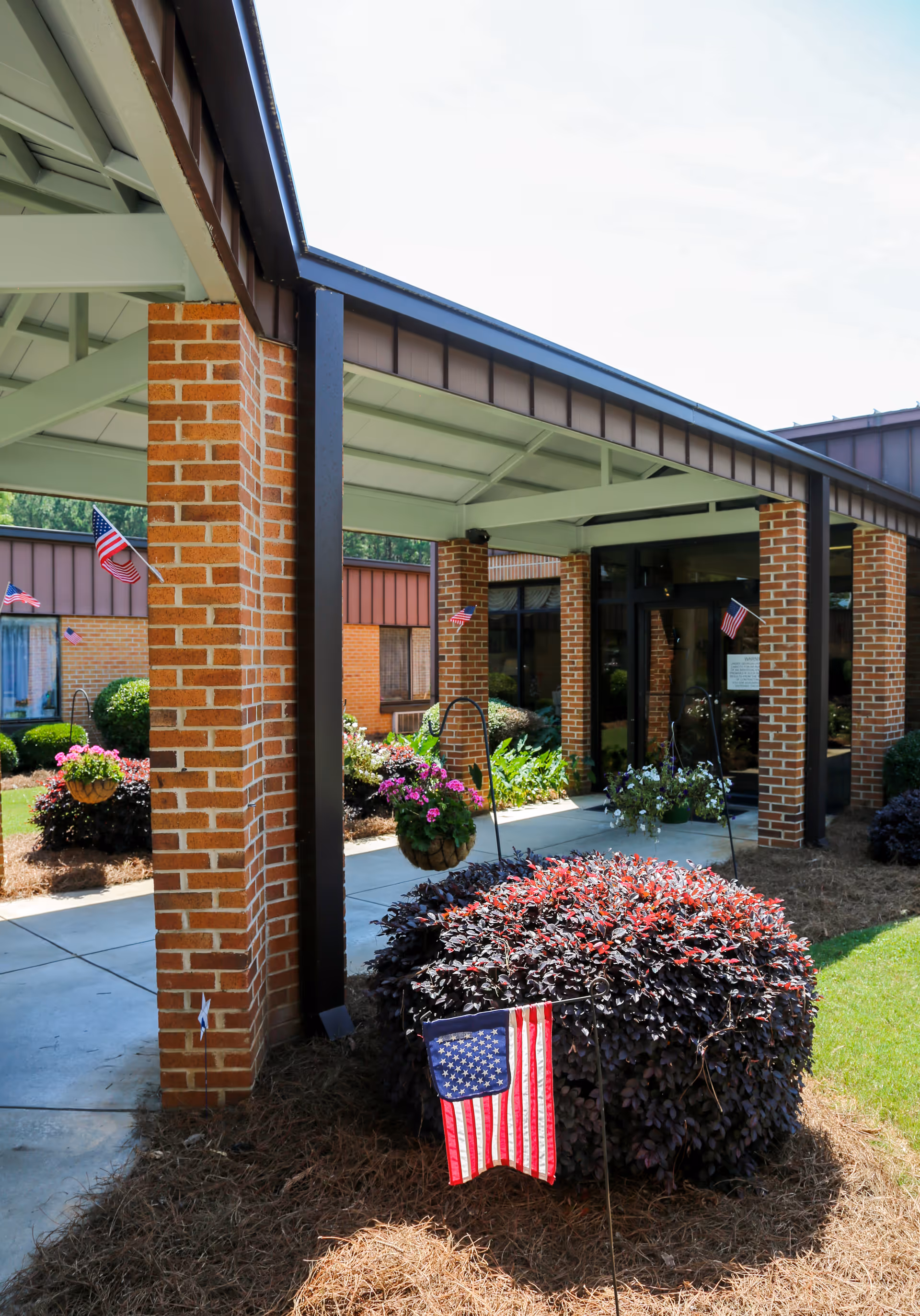 Covered entrance area of a brick building with hanging flower baskets and American flags displayed in the landscaping. The entrance has a roof supported by brick columns and a glass door.