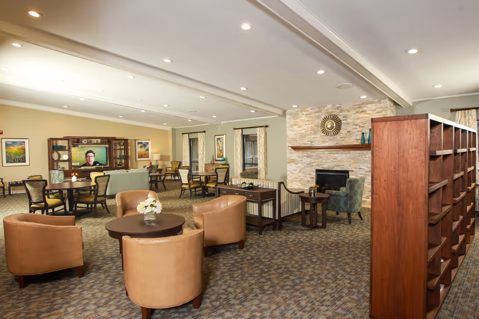 Spacious lounge with armchairs, tables, a fireplace, and a TV in a senior living facility.