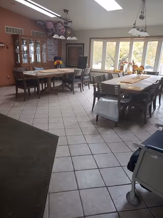 Spacious dining room with two long tables and chairs, a china cabinet, large windows, tiled floor and overhead lights.