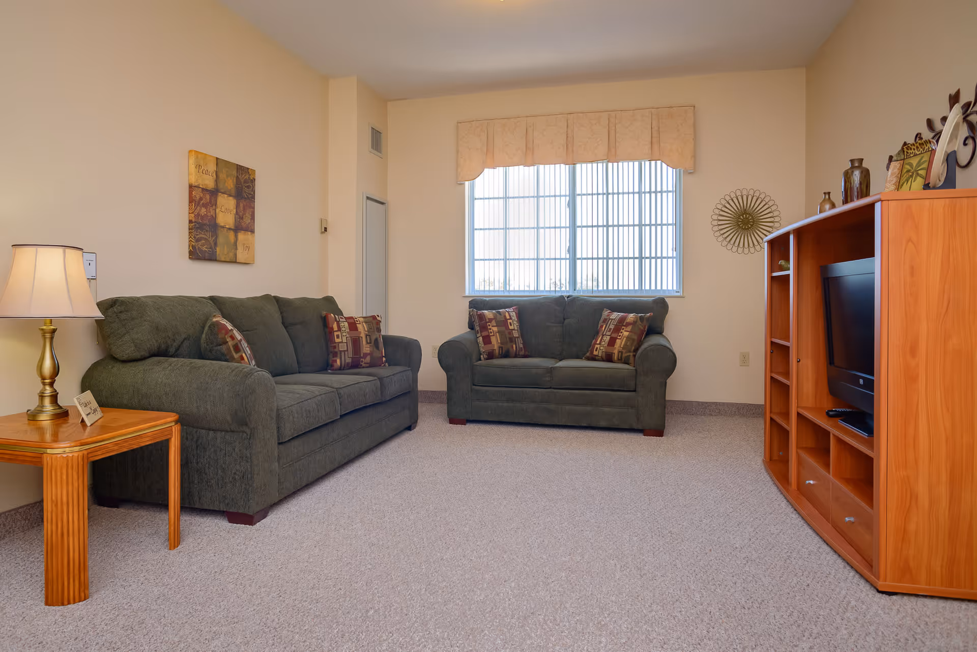 A cozy living room with two green upholstered sofas, each adorned with patterned throw pillows. A wooden side table with a lamp and a small decorative sign sits next to one sofa. Across from the sofas is a wooden entertainment center holding a flat-screen TV and decorative items. A window with vertical blinds and a beige valance lets in natural light, and the walls are painted a soft cream color.