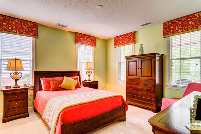 A bright bedroom with light green walls and three windows with red floral valances. The room features a wooden bed with a red bedspread, red and yellow pillows, two wooden nightstands with lamps, a wooden dresser with a decorative vase on top, a pink armchair, and a wooden desk with a mirror.