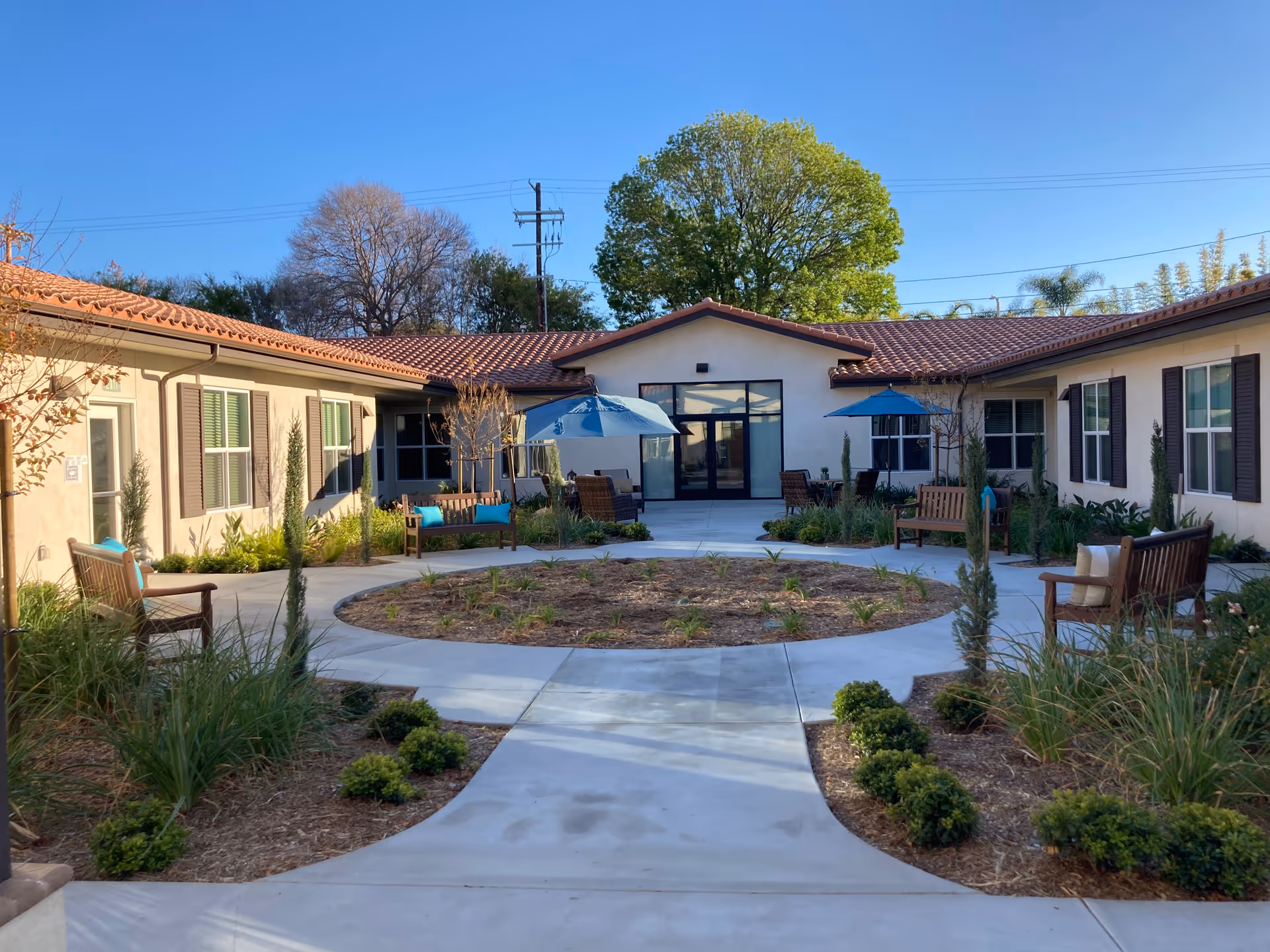 Outdoor courtyard area of The Preserve at Woodland Hills Assisted Living & Memory Care featuring a circular garden bed in the center, surrounded by a concrete walkway, benches with cushions, patio tables with blue umbrellas, and landscaped plants. The building has beige walls, brown shutters, and a red tile roof under a clear blue sky.