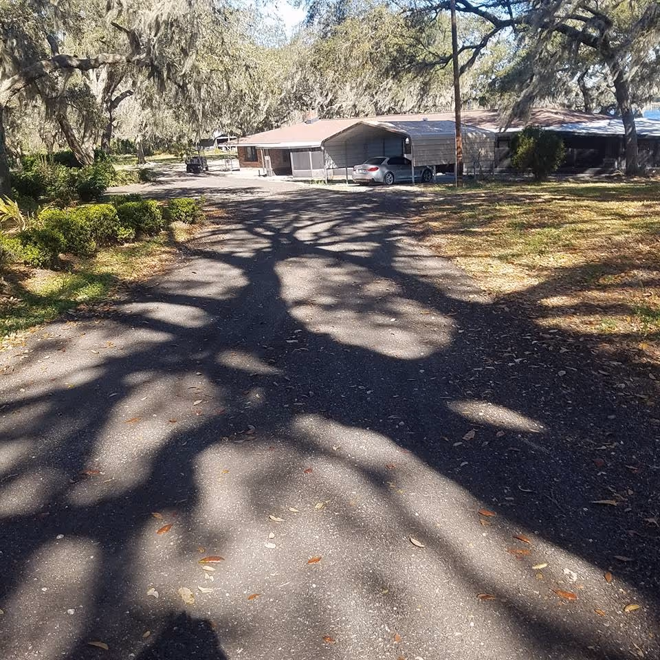 A paved driveway leading to a single-story building with a carport and a silver car parked underneath. Large trees with hanging moss cast shadows on the driveway and surrounding grassy areas with bushes.