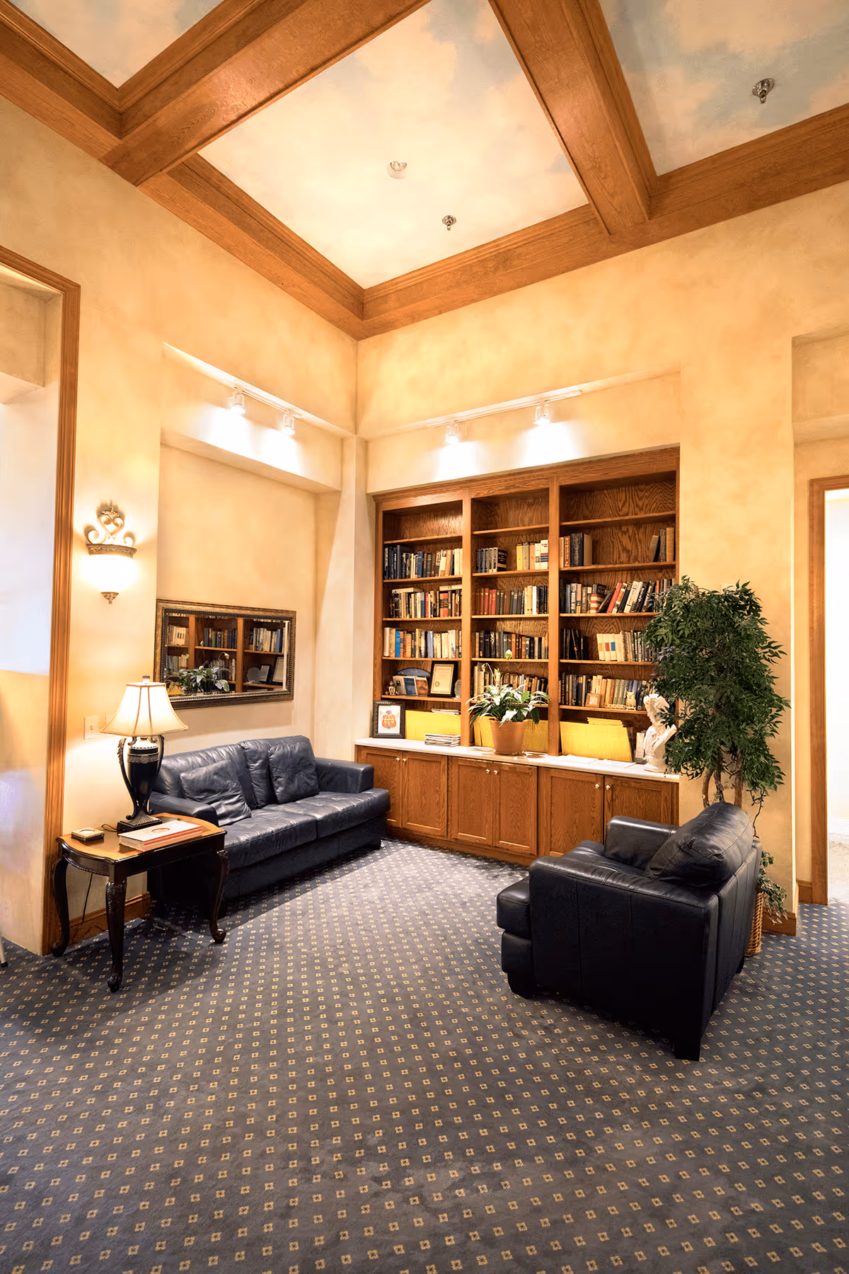 A cozy living room area with two black leather sofas, a wooden side table with a lamp, a large built-in wooden bookshelf filled with books and decorative items, a potted plant, and a ceiling with wooden beams and a sky mural.