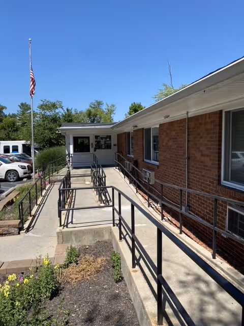 Exterior view of a brick building with a wheelchair accessible ramp leading to the entrance. There is a flagpole with an American flag on the left side, some parked cars, and a small garden area with plants in the foreground. The sky is clear and blue.