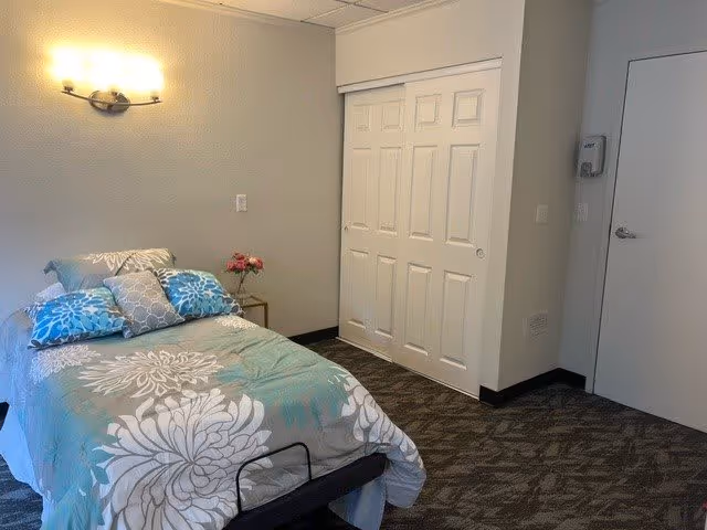 A small furnished bedroom with a single bed covered in floral bedding, a wall sconce, closet doors, and a side table with flowers.
