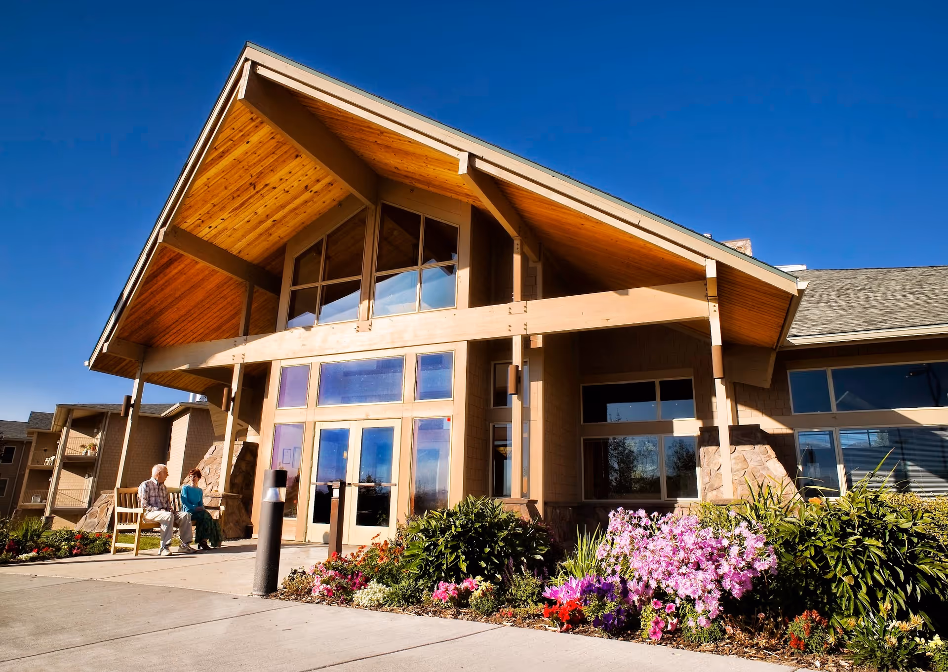 Exterior view of Bozeman Health Hillcrest Senior Living, Aspen Pointe building with a wooden peaked roof, large glass windows, and a flower bed with colorful flowers in front. Two elderly people are sitting on a bench near the entrance under a clear blue sky.