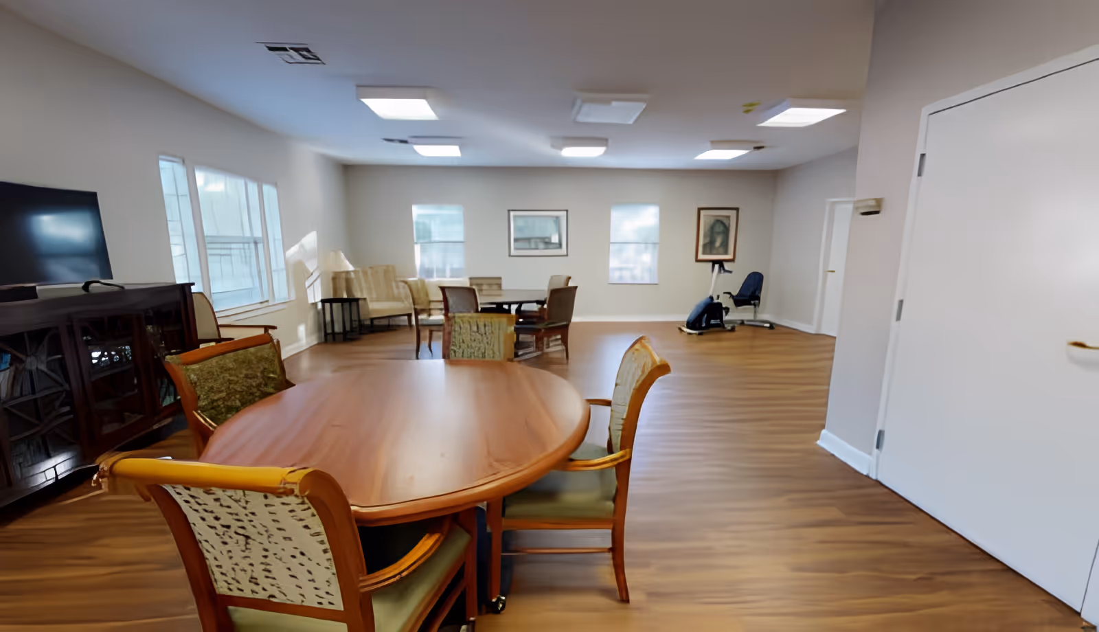 A spacious senior living common area with wooden floors, multiple tables and chairs, a TV on a wooden cabinet, large windows, framed artwork on the walls, and an exercise bike in the corner.
