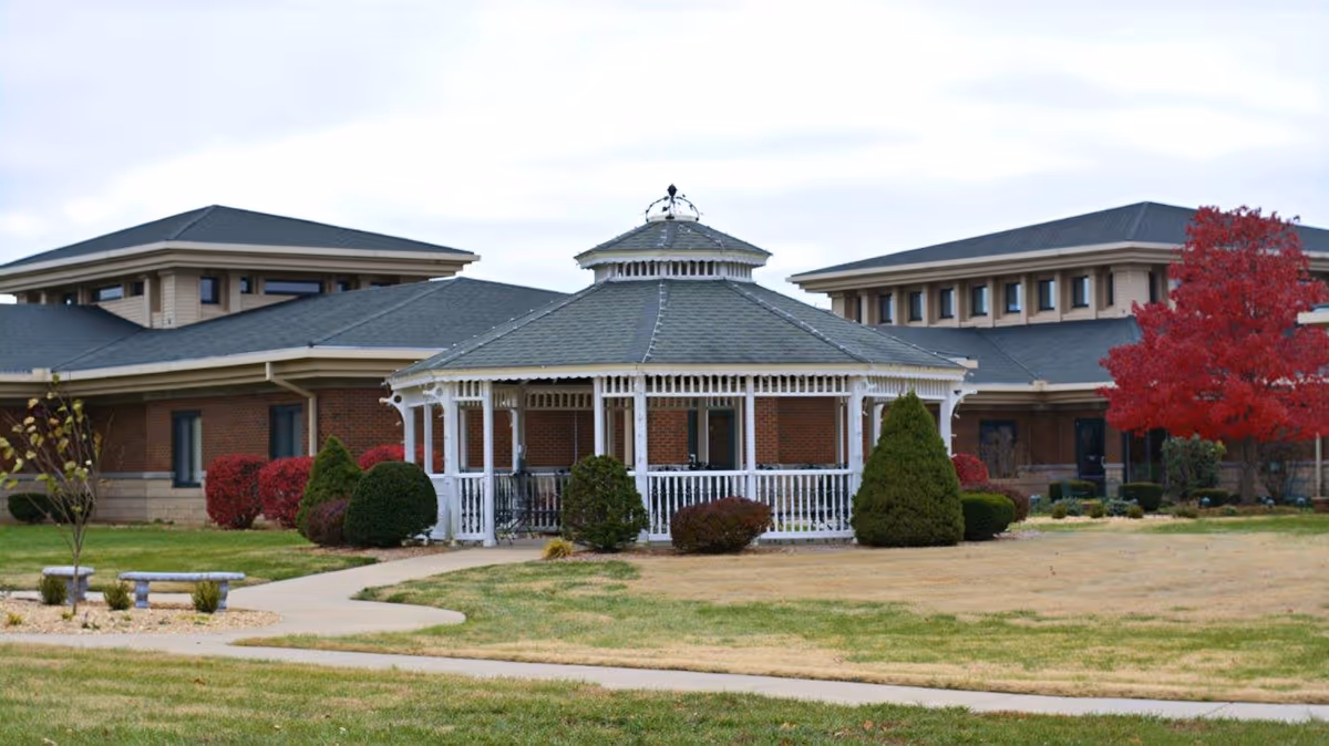 Outdoor view of a senior living facility with a white gazebo in the center surrounded by trimmed bushes and a paved walkway. The building in the background has a brick exterior with multiple windows and a dark roof. There is a red tree on the right side of the image.