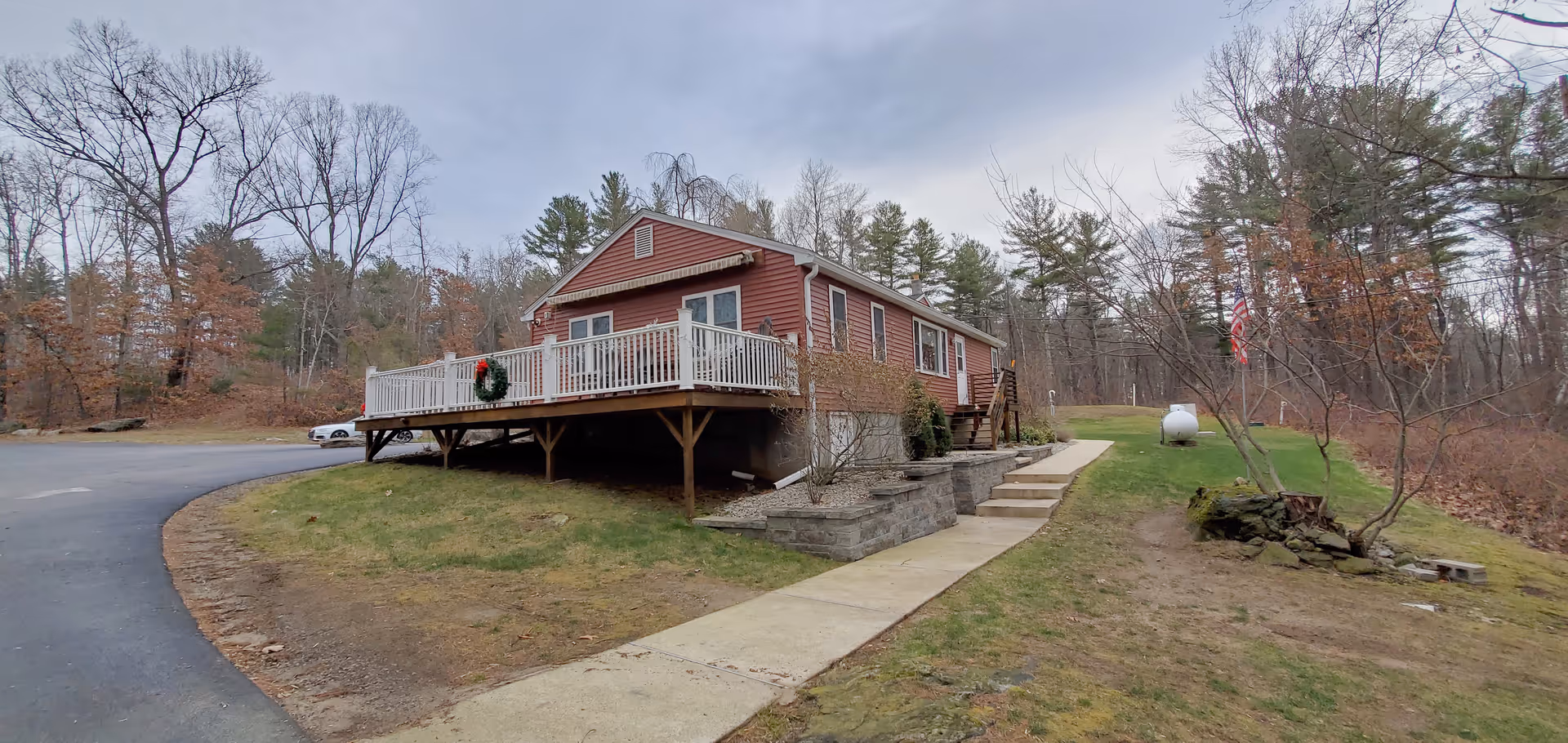 A single-story red building with white railings on a raised wooden deck, surrounded by a grassy area and trees with bare branches. There is a concrete walkway leading to the building, and an American flag is visible on the right side near a propane tank. The sky is overcast.