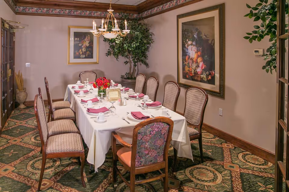 A formal dining room with a long table set for a meal, surrounded by upholstered chairs, a chandelier, framed artwork, and a floral centerpiece.