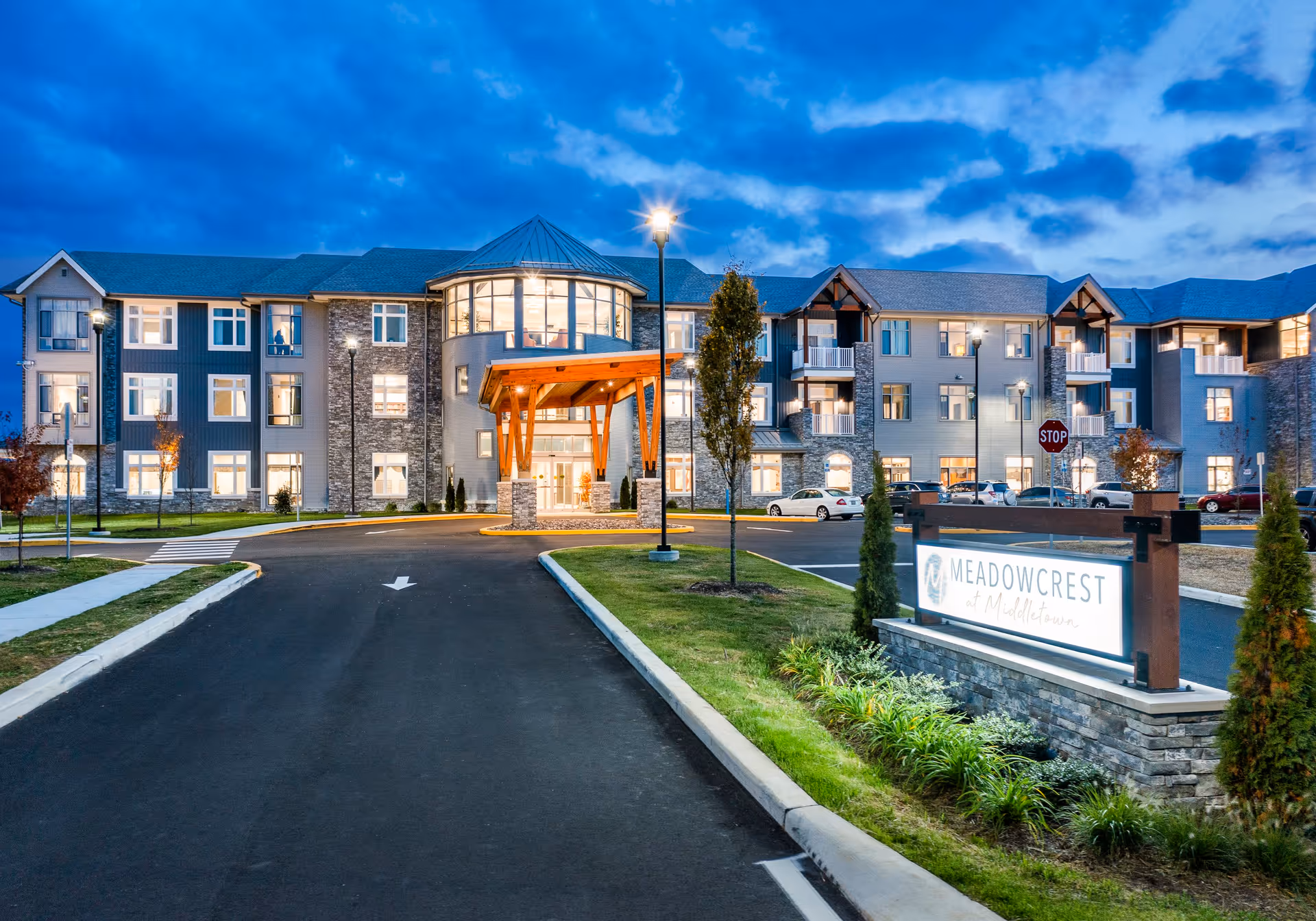 Exterior view of Meadowcrest at Middletown senior living facility at dusk, showing a large three-story building with illuminated windows, a covered entrance with wooden beams, a driveway leading to the entrance, landscaped greenery, and a sign with the facility's name.