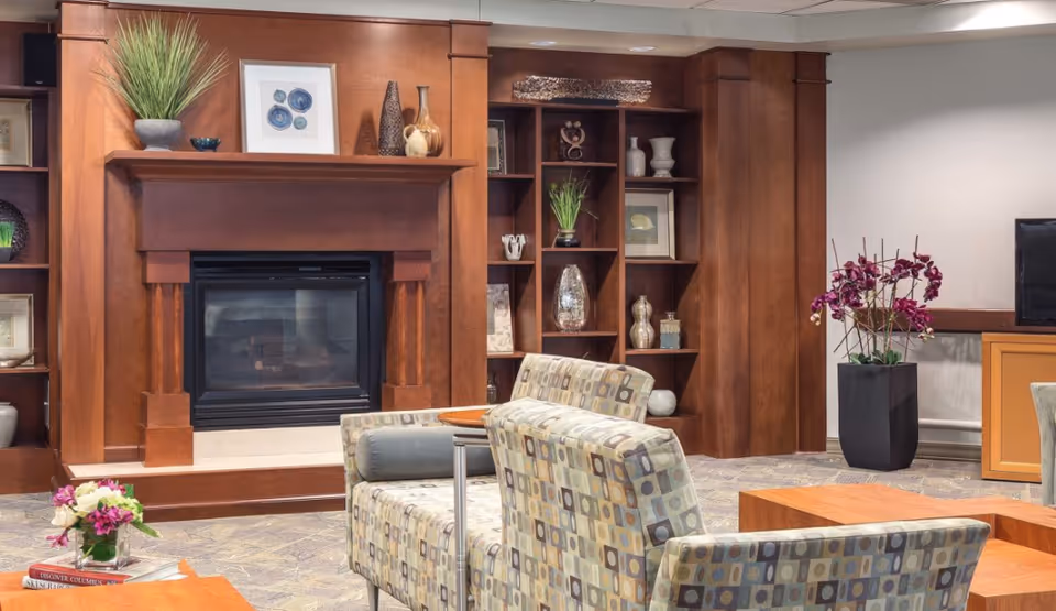 A cozy living room area with patterned armchairs, a wooden fireplace mantel decorated with vases and framed art, built-in wooden shelves with decorative items, a flat-screen TV on a wooden stand, and a tall black planter with purple flowers.