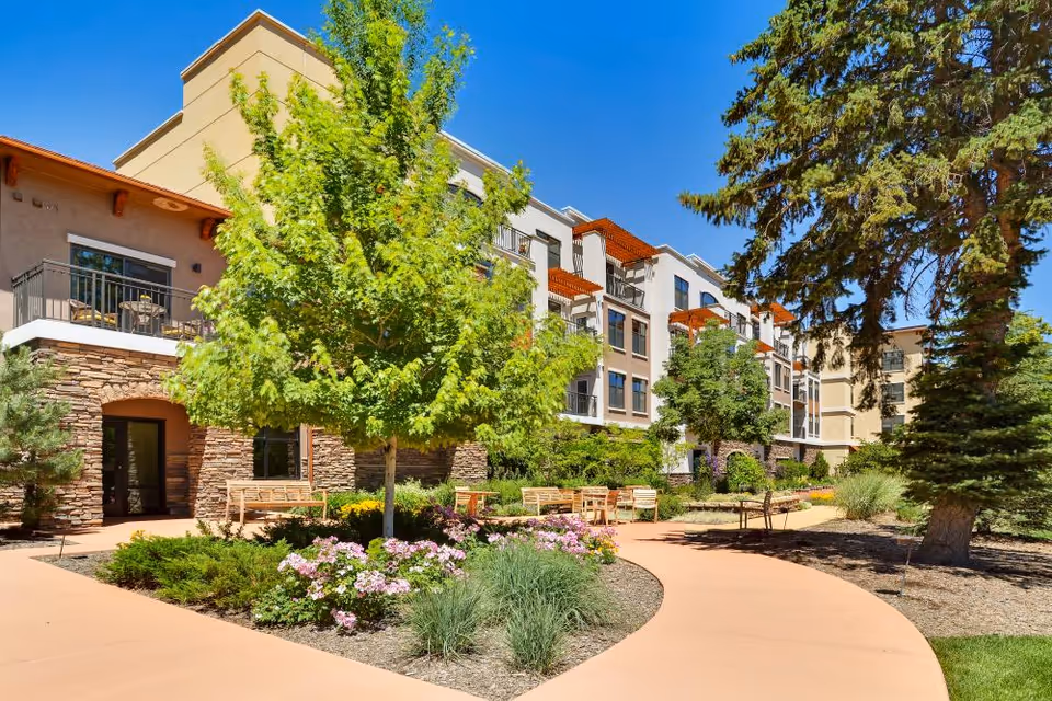 Sunlit courtyard with a curved paved path, benches, trees and flower beds in front of a multi-story senior living building.
