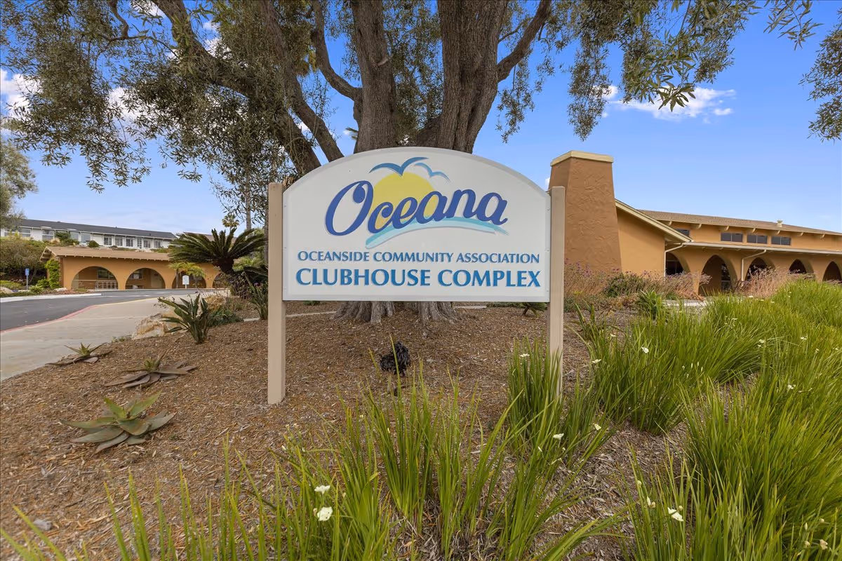 A large sign in front of a clubhouse complex reads 'Oceana Oceanside Community Association Clubhouse Complex' with a tree and landscaped plants around it. The clubhouse building with arches is visible in the background under a blue sky.
