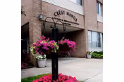 Entrance of Crest Manor Living & Rehab Center with a brick facade, hanging flower baskets, and a sidewalk.