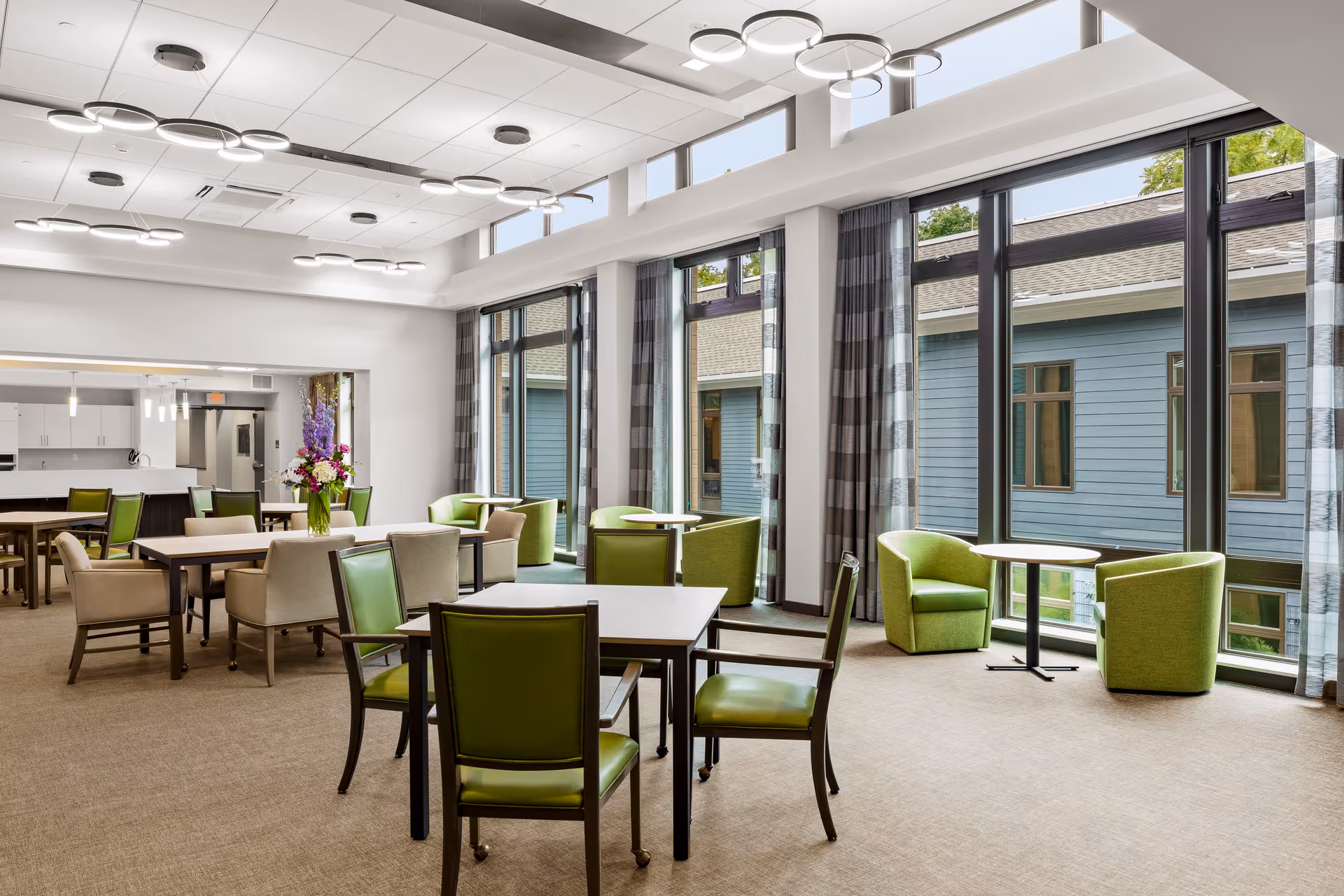 A bright and spacious dining area with multiple tables and chairs. The chairs are a mix of green and beige upholstery. Large floor-to-ceiling windows with gray plaid curtains let in natural light and offer a view of an adjacent building with blue siding. Modern circular ceiling lights illuminate the room, and a vase with purple and white flowers is placed on one of the tables.